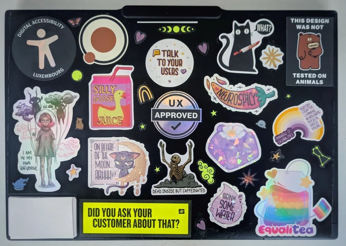 A laptop lid covered with colorful stickers, including digital accessibility, UX approved, talk to your users, tested on animals, and equalitea (not a typo, a tea bag a rainbow) a fun one with a skeletons that reads dead inside but caffeinated, one that reads did you ask your customers about that, and the sailor moon fan saying On behalf of the moon argh. Also a drink some water one, a silly goose juice one, a neuropsicy one, and a cat with a knife going what!?